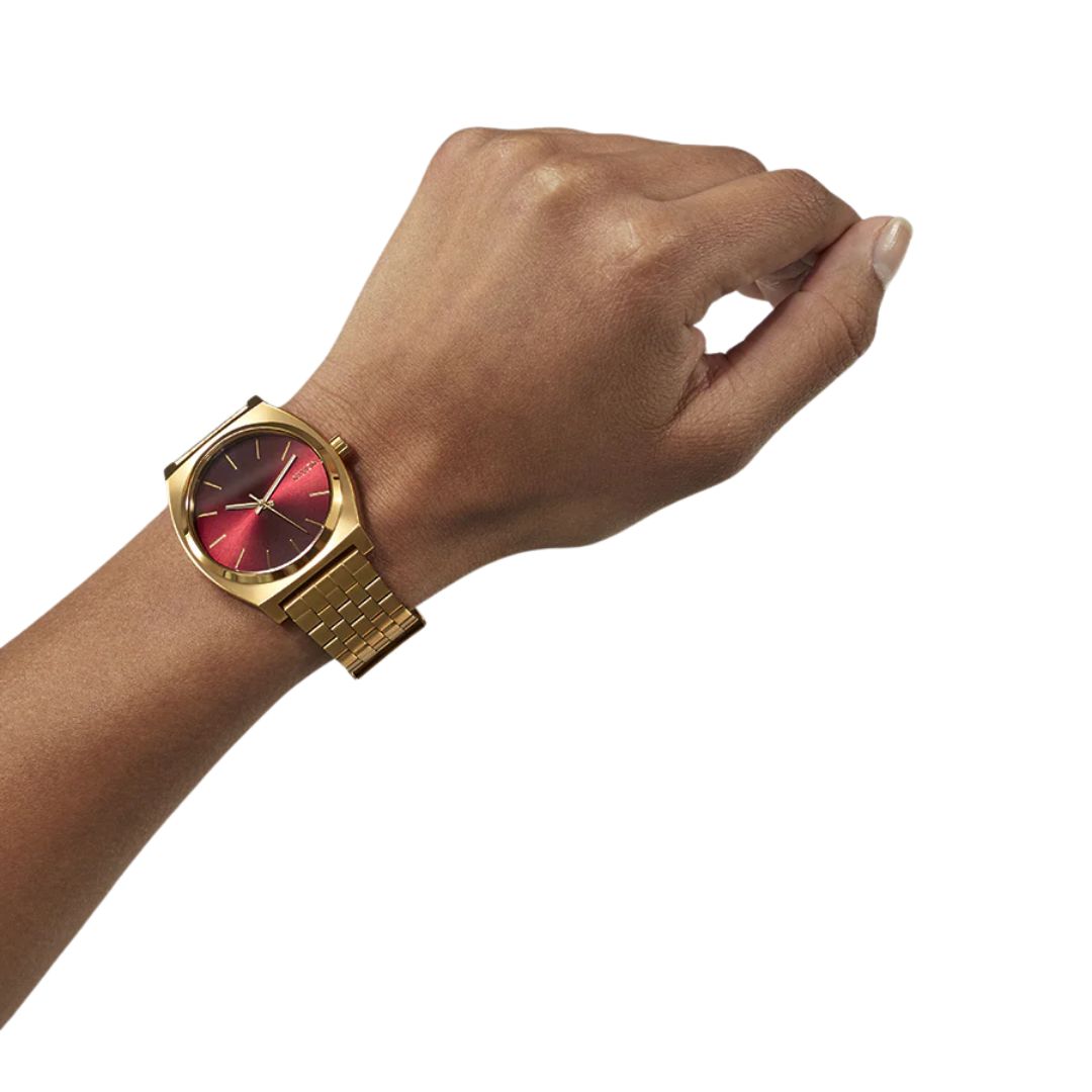 Reloj Nixon Time Teller dorado con esfera rojo oxblood sunray en muñeca masculina.