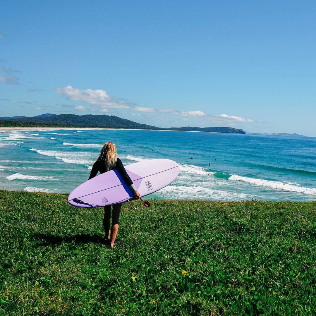 Surfista caminando hacia el mar con una tabla de surf softboard Ocean & Earth Happy Hour Epoxy Soft bajo el brazo, escena en costa australiana.