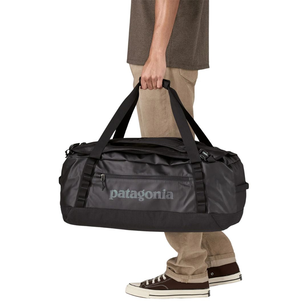 Patagonia Black Hole Duffel 40L en color black on black, vista lateral con modelo sujetándola por las asas reforzadas de nailon reciclado, mostrando el bolsillo frontal con cremallera, el logo Patagonia en tono grisáceo sobre tejido ripstop negro mate laminado con TPU reciclado y los puntos de sujeción exteriores.