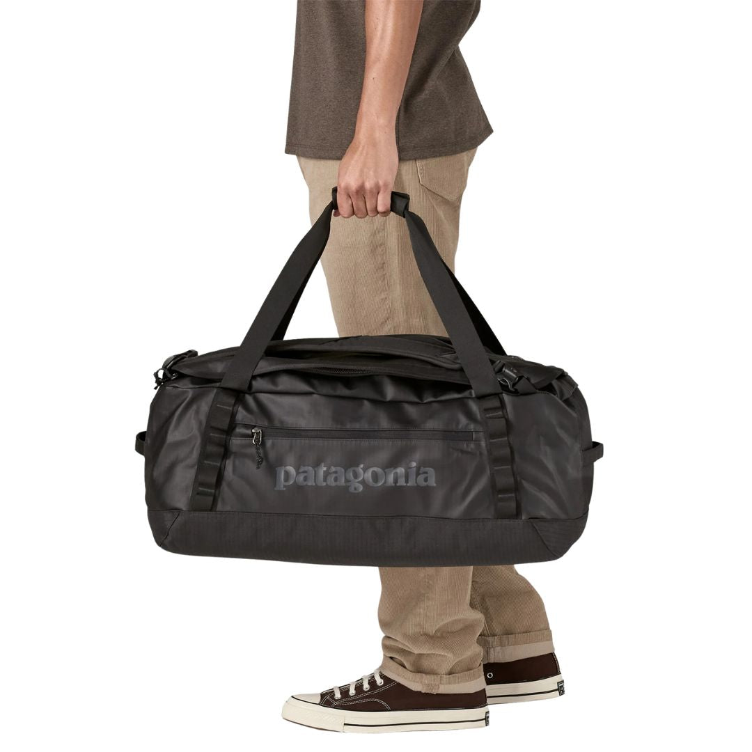 Patagonia Black Hole Duffel 40L en color black on black, vista frontal con modelo transportándola por las asas unidas, mostrando el volumen de 40 litros, el bolsillo lateral con cremallera, el logo Patagonia en acabado mate sobre ripstop negro laminado con TPU y los puntos de sujeción de cadena margarita.