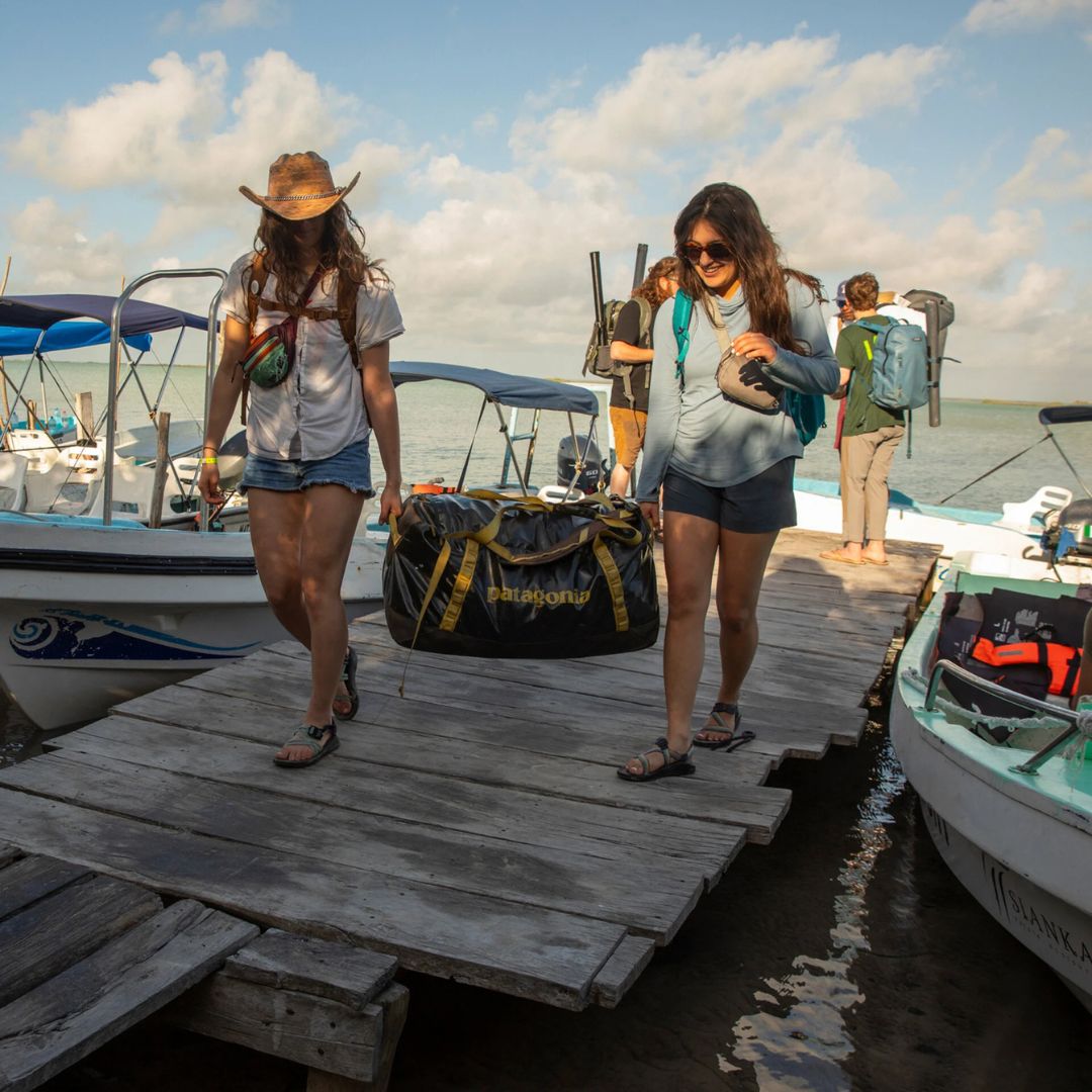 Patagonia Black Hole Duffel 40L en color negro con correas doradas, transportada entre dos personas por las asas reforzadas en un embarcadero con barcos al fondo, mostrando la resistencia y versatilidad de la bolsa de viaje en entorno marítimo y outdoor.