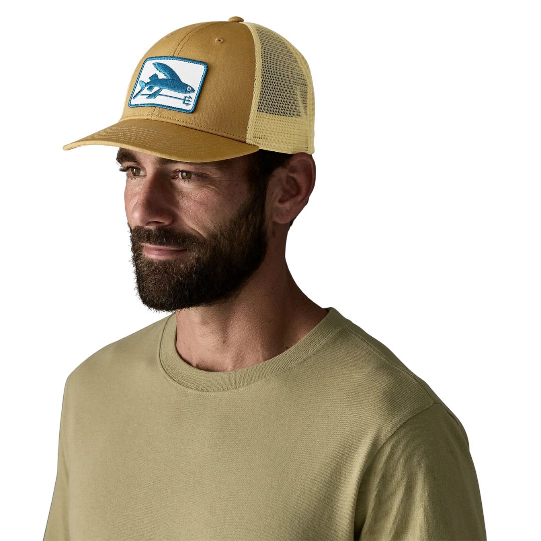 Hombre con gorra trucker Patagonia Flying Fish LoPro en color Bobcat Brown, vista frontal, con parche bordado de pez volador en azul sobre fondo blanco y parte trasera de malla beige.