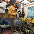 Chaqueta polar Patagonia Better Sweater en tono mostaza, hombre guardando unas botas de montaña en un macuto Patagonia entre bolsas de viaje, mostrando la cremallera completa y el bolsillo de pecho.