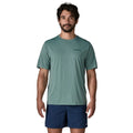 Patagonia Men's Capilene Cool Daily Shirt Boardshort Logo en color blue sage, vista frontal con modelo combinada con bañador azul marino, camiseta técnica de manga corta con logo Patagonia discreto en el pecho izquierdo sobre tejido de poliéster reciclado jaspeado verde azulado.