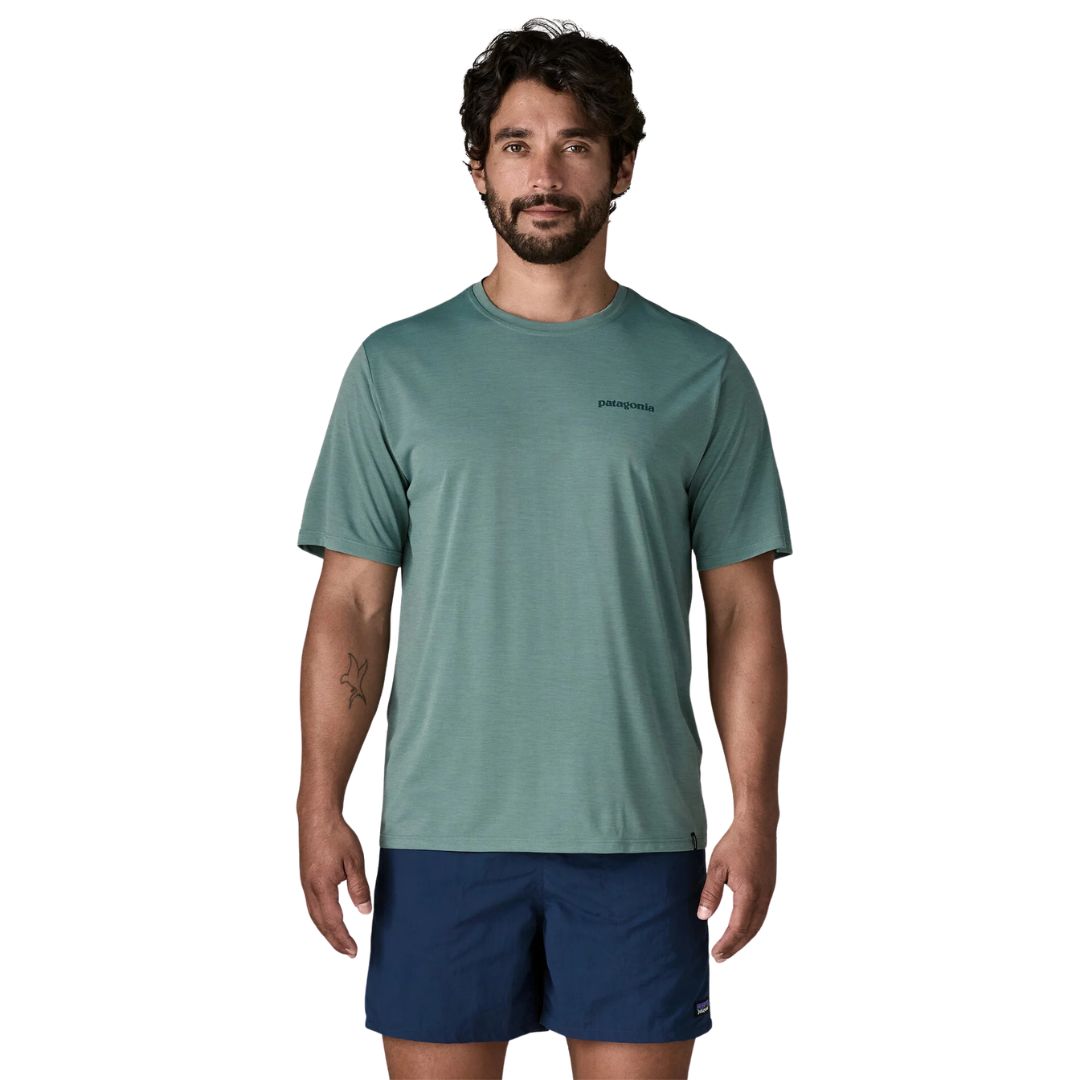 Patagonia Men's Capilene Cool Daily Shirt Boardshort Logo en color blue sage, vista frontal con modelo combinada con bañador azul marino, camiseta técnica de manga corta con logo Patagonia discreto en el pecho izquierdo sobre tejido de poliéster reciclado jaspeado verde azulado.
