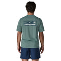 Patagonia Men's Capilene Cool Daily Shirt Boardshort Logo en color blue sage, vista trasera con modelo, mostrando el estampado icónico Boardshort Logo con olas y surf en tonos verde petróleo y crema sobre fondo verde azulado jaspeado.
