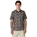 Patagonia Men's Go-To Shirt en estampado jaguar geo dolomite blue, vista frontal con modelo combinada con pantalón corto beige, mostrando el cuello con solapas, la tapeta de botones, el bolsillo izquierdo del pecho y el estampado geométrico de triángulos con palmeras y jaguares en tonos azul marino oscuro, beige rosado y verde oliva sobre tejido ligero de algodón orgánico y poliéster reciclado.