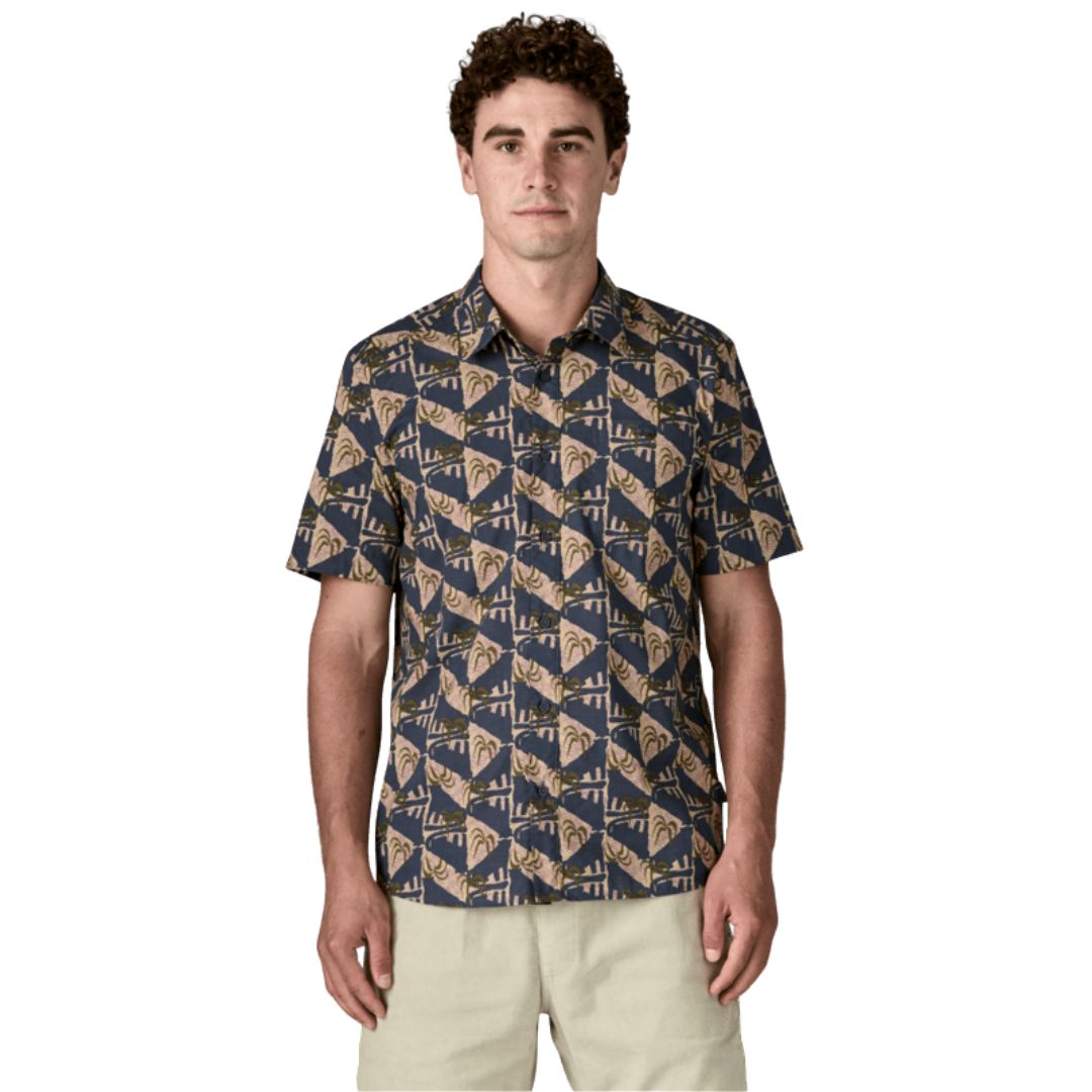 Patagonia Men's Go-To Shirt en estampado jaguar geo dolomite blue, vista frontal con modelo combinada con pantalón corto beige, mostrando el cuello con solapas, la tapeta de botones, el bolsillo izquierdo del pecho y el estampado geométrico de triángulos con palmeras y jaguares en tonos azul marino oscuro, beige rosado y verde oliva sobre tejido ligero de algodón orgánico y poliéster reciclado.