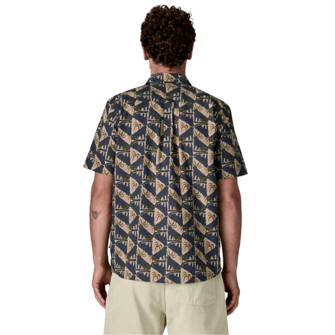Patagonia Men's Go-To Shirt en estampado jaguar geo dolomite blue, vista trasera con modelo combinada con pantalón corto beige arena, mostrando el estampado geométrico de triángulos alternados en azul marino oscuro y beige rosado con motivos de palmeras y jaguares en verde oliva, el bajo redondeado y la construcción del tejido de algodón orgánico y poliéster reciclado.