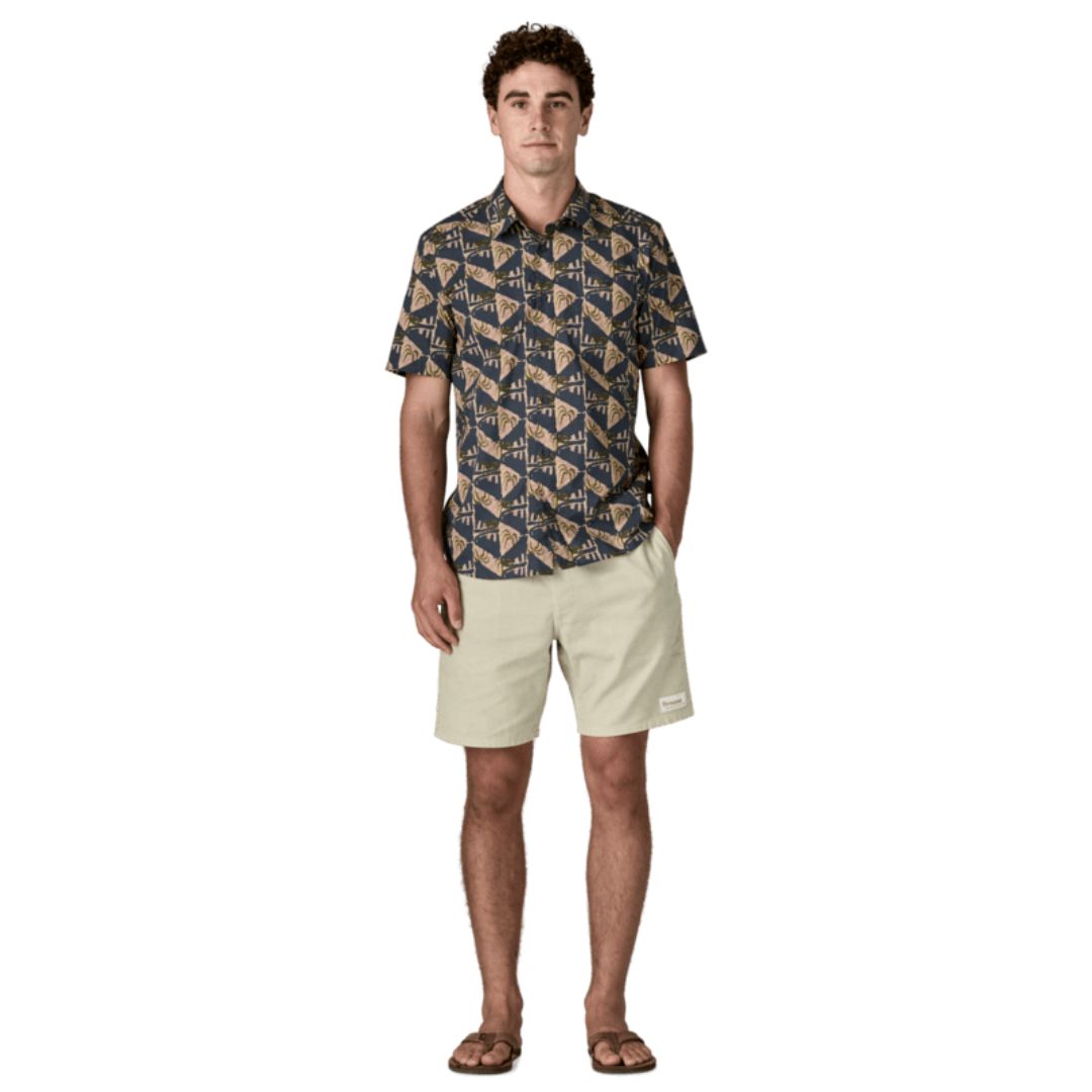 Patagonia Men's Go-To Shirt en estampado jaguar geo dolomite blue, vista frontal completa con modelo combinada con pantalón corto beige arena y chanclas marrones, mostrando el corte slim fit, el estampado geométrico de triángulos con motivos de palmeras y jaguares en azul marino y beige, el bolsillo izquierdo del pecho y el bajo redondeado por fuera del pantalón.