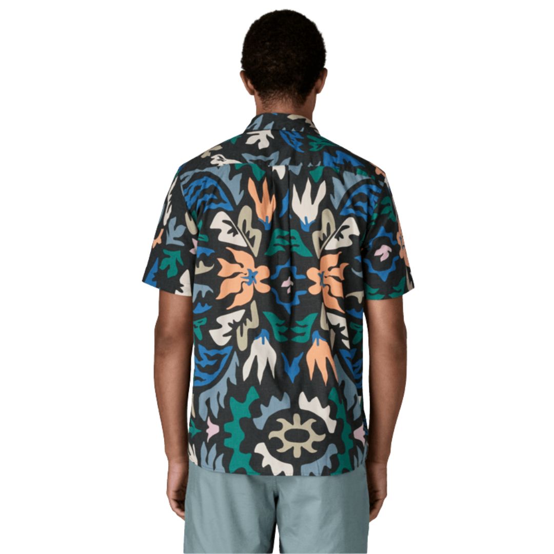 Patagonia Men's Go-To Shirt en estampado kaleido black, vista trasera con modelo combinada con pantalón corto azul grisáceo, mostrando el estampado caleidoscópico de flores y formas orgánicas en tonos naranja, verde, azul, crema y negro sobre tejido de algodón orgánico y poliéster reciclado, y el bajo redondeado.