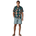 Patagonia Men's Go-To Shirt en estampado kaleido black, vista frontal completa con modelo combinada con pantalón corto azul grisáceo y chanclas, mostrando el corte slim fit, el estampado caleidoscópico multicolor sobre fondo negro con flores en naranja, verde y azul, y el bajo redondeado por fuera del pantalón.