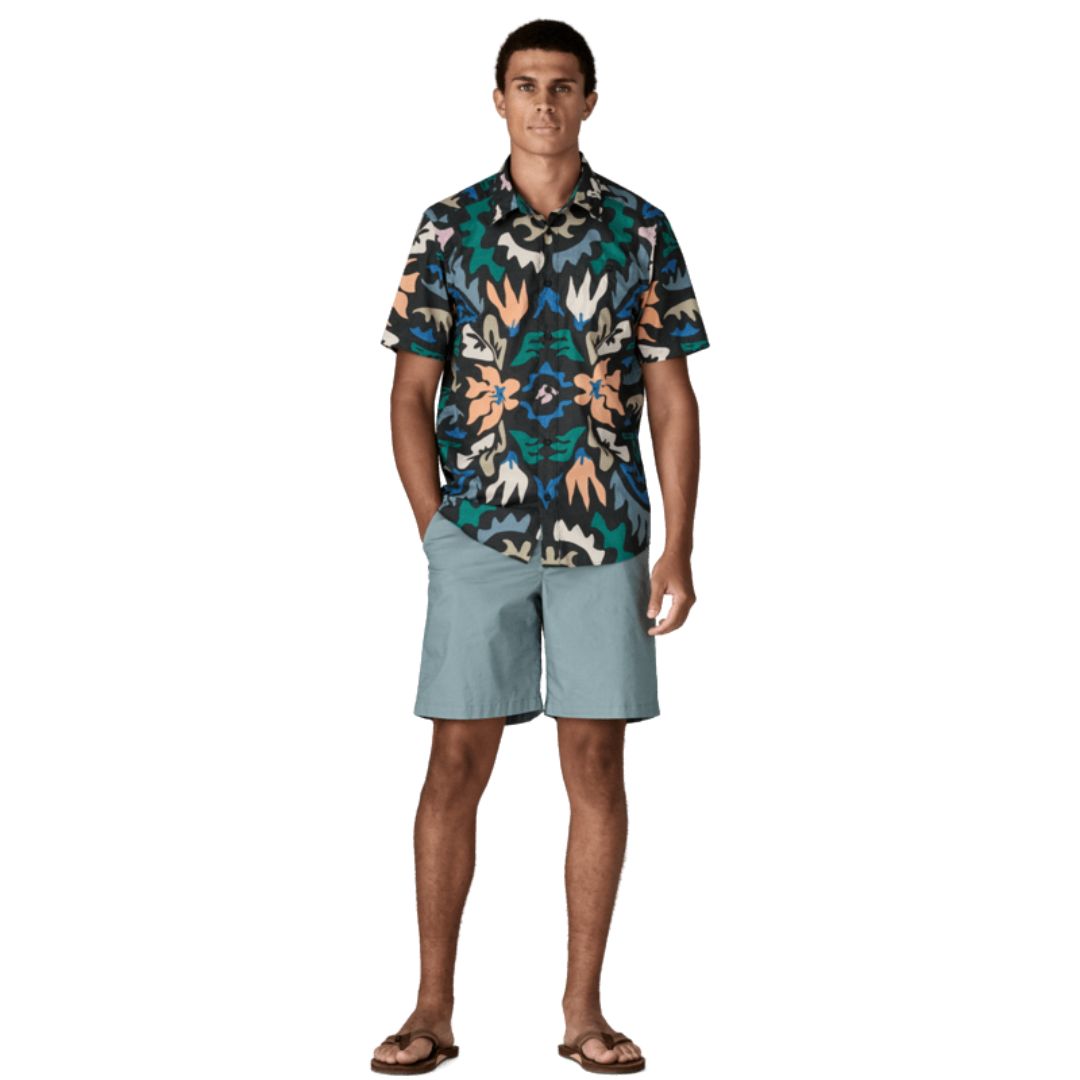 Patagonia Men's Go-To Shirt en estampado kaleido black, vista frontal completa con modelo combinada con pantalón corto azul grisáceo y chanclas, mostrando el corte slim fit, el estampado caleidoscópico multicolor sobre fondo negro con flores en naranja, verde y azul, y el bajo redondeado por fuera del pantalón.