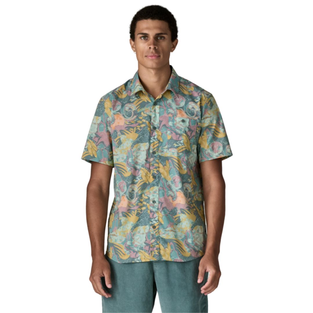 Patagonia Men's Go-To Shirt en estampado moon tripper blue sage, vista frontal con modelo combinada con pantalón corto verde azulado, mostrando el cuello con solapas, la tapeta de botones, el bolsillo izquierdo del pecho y el estampado tropical de figuras lunares y hojas en tonos verde azulado, amarillo mostaza y rosa sobre tejido ligero de algodón orgánico y poliéster reciclado.