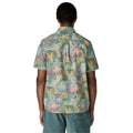 Patagonia Men's Go-To Shirt en estampado moon tripper blue sage, vista trasera con modelo combinada con pantalón corto azul verdoso, mostrando el estampado tropical de figuras lunares, hojas y formas orgánicas en tonos verde azulado, amarillo mostaza, rosa y melocotón sobre tejido de algodón orgánico y poliéster reciclado, y el bajo redondeado.