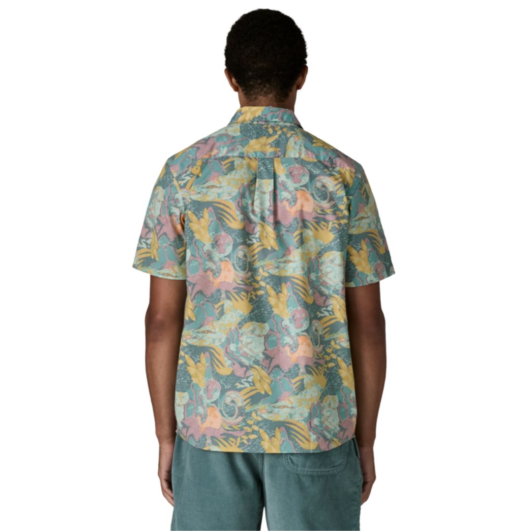 Patagonia Men's Go-To Shirt en estampado moon tripper blue sage, vista trasera con modelo combinada con pantalón corto azul verdoso, mostrando el estampado tropical de figuras lunares, hojas y formas orgánicas en tonos verde azulado, amarillo mostaza, rosa y melocotón sobre tejido de algodón orgánico y poliéster reciclado, y el bajo redondeado.