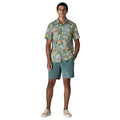 Patagonia Men's Go-To Shirt en estampado moon tripper blue sage, vista frontal completa con modelo combinada con pantalón corto verde azulado y zapatillas blancas, mostrando el corte slim fit abierto, el estampado tropical multicolor en tonos verde azulado, amarillo mostaza, rosa y melocotón, el bolsillo izquierdo del pecho y el bajo redondeado por fuera del pantalón.
