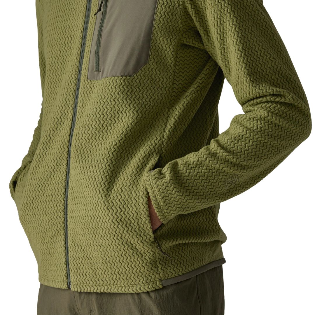 Patagonia Men's R1 Air Fleece Jacket en color caper green, detalle lateral del bajo con modelo usando el bolsillo delantero con cremallera minimalista, tejido jacquard en zigzag y ribete tejido en el bajo.