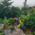 Grupo de senderistas con chaquetas impermeables Patagonia Torrentshell 3L ascendiendo terreno rocoso bajo lluvia y niebla con mochilas de montaña.