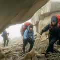 Senderistas con chaquetas impermeables Patagonia ascendiendo terreno rocoso en condiciones de lluvia intensa, niebla y frío con bastones y mochilas de alta montaña.