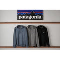 Sudadera con capucha Patagonia P-6 Label Uprisal Hoody colgada en gris Gravel Heather junto a otros colores de la gama, bajo el rótulo de montañas Patagonia sobre pared blanca.