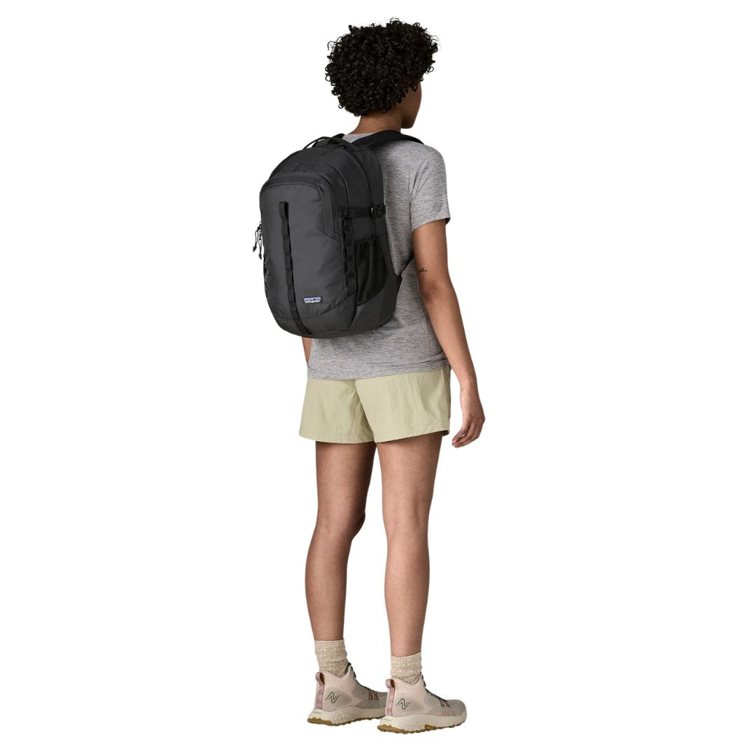 Patagonia Refugio Daypack 26L en color negro, vista trasera completa con modelo en contexto outdoor, mostrando el volumen de la mochila, el bolsillo frontal con cremallera, el bolsillo lateral de malla elástica y el panel trasero transpirable.