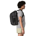 Patagonia Refugio Daypack 26L en color negro, vista lateral con modelo, mostrando el perfil de la mochila, el bolsillo lateral de malla elástica, la correa esternal y el asa superior de transporte.