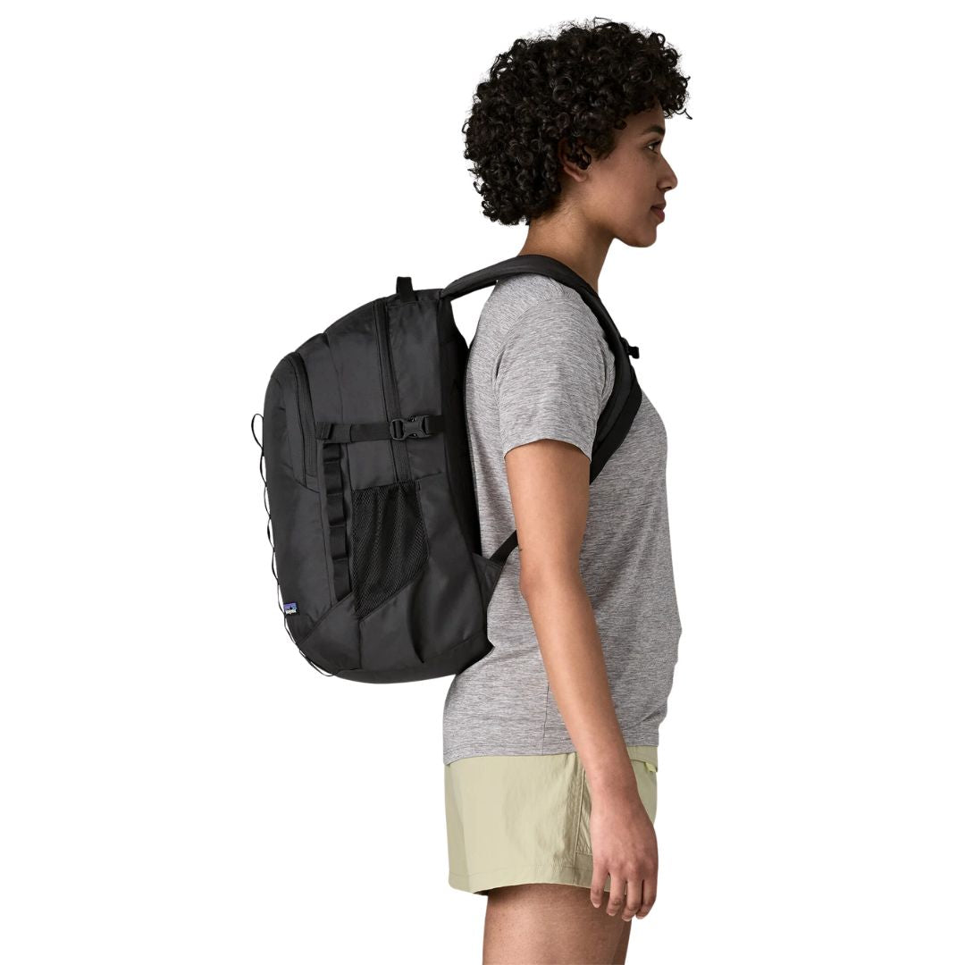 Patagonia Refugio Daypack 26L en color negro, vista lateral con modelo, mostrando el perfil de la mochila, el bolsillo lateral de malla elástica, la correa esternal y el asa superior de transporte.