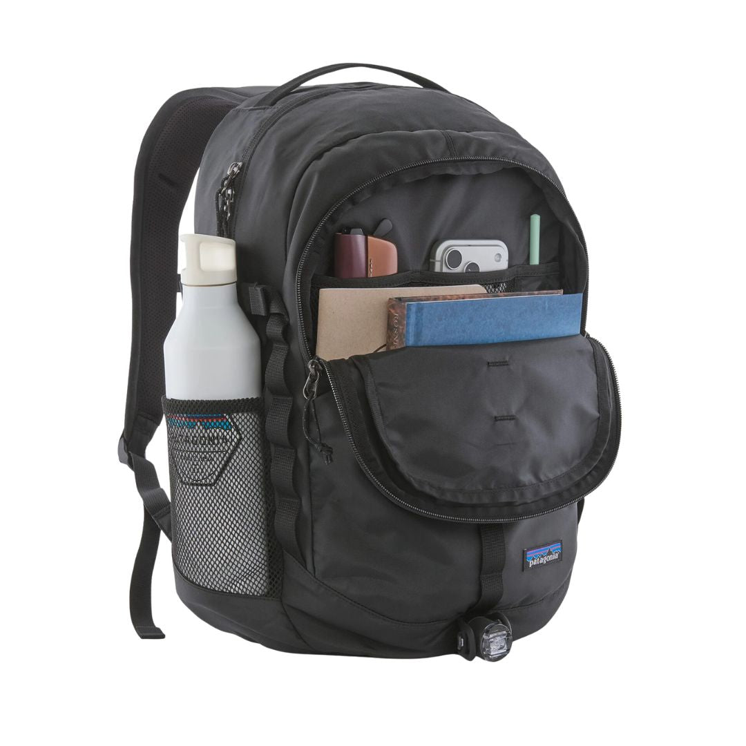 Patagonia Refugio Daypack 26L en color negro, compartimento frontal abierto con organización interna mostrando cuadernos, móvil y accesorios, bolsillo lateral de malla elástica con botella de agua y luz de bicicleta enganchada en la base.