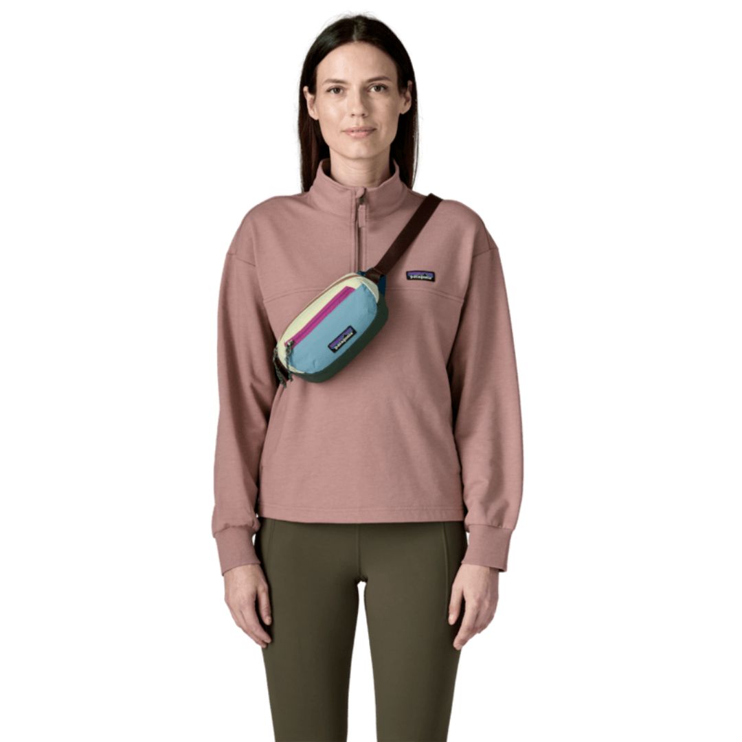 Riñonera Patagonia Terravia Mini Hip Pack en colorway Patchwork Snowmelt Blue, modelo llevándola cruzada sobre el pecho a modo de bandolera sobre una sudadera rosa.