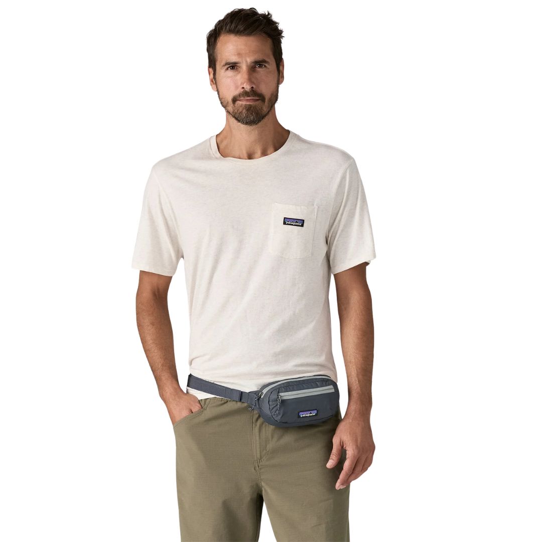 Patagonia Terravia Mini Hip Pack 1L en color smolder blue, vista frontal completa con modelo llevándola como riñonera en la cintura, mostrando el compartimento principal con franja gris, el bolsillo frontal de seguridad, la etiqueta Patagonia y el cinturón antideslizante azul marino.