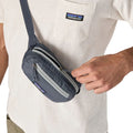 Patagonia Terravia Mini Hip Pack 1L en color smolder blue, detalle de acceso al bolsillo frontal de seguridad con cremallera y tirador azul marino, llevada como bolso cruzado, mostrando la doble cremallera y el tejido ripstop de nailon reciclado resistente a la intemperie.