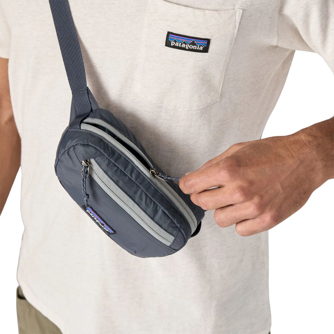 Patagonia Terravia Mini Hip Pack 1L en color smolder blue, detalle de acceso al bolsillo frontal de seguridad con cremallera y tirador azul marino, llevada como bolso cruzado, mostrando la doble cremallera y el tejido ripstop de nailon reciclado resistente a la intemperie.