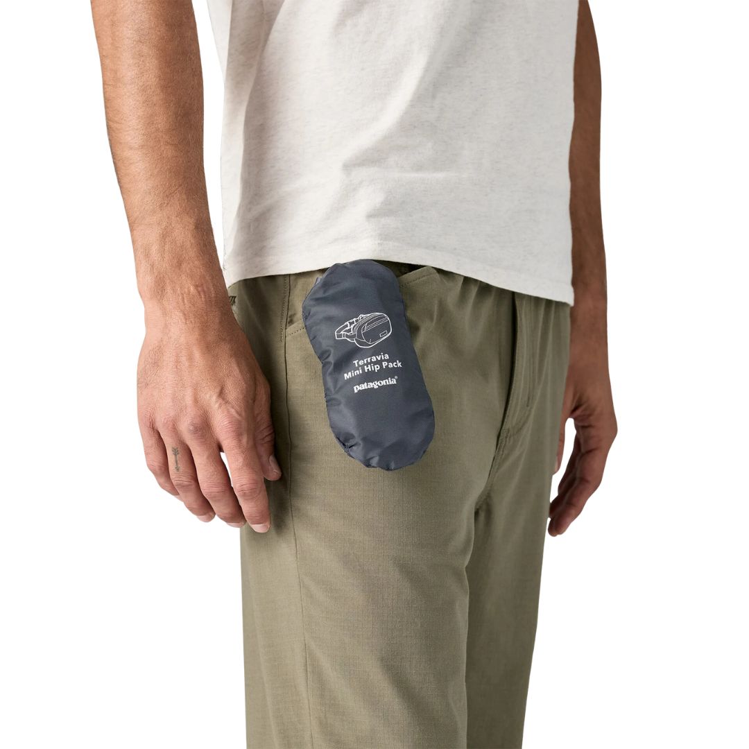 Patagonia Terravia Mini Hip Pack 1L en color smolder blue plegada en su propio bolsillo interior, con el texto Terravia Mini Hip Pack y el logo Patagonia visibles, guardada en el bolsillo lateral del pantalón del modelo para demostrar su diseño ultracompacto y plegable.