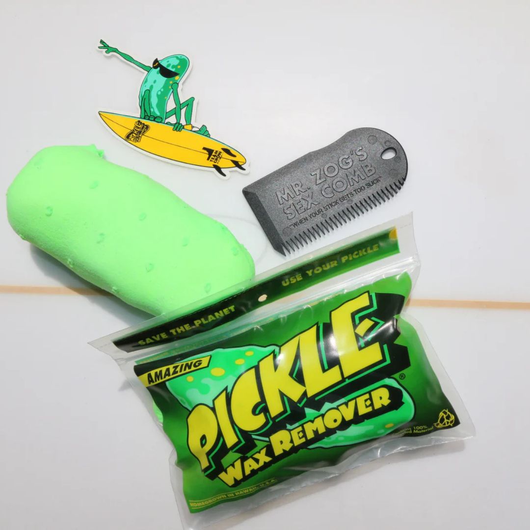 Pickle Wax Remover pepino mágico de Team Chow en verde lima sobre tabla de surf, junto a peine rascador gris y bolsa biodegradable resellable con pegatina ilustrada