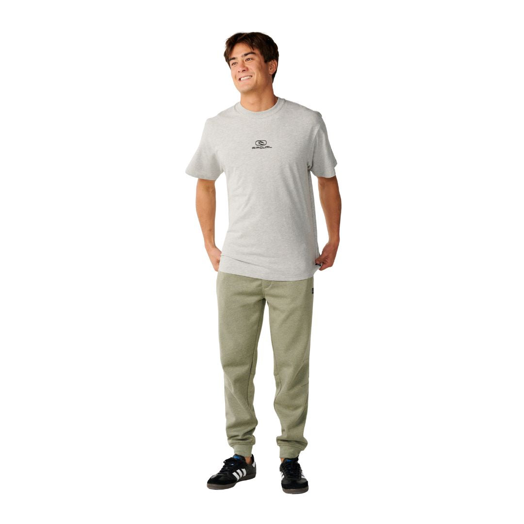 Rip Curl Anti-Series Departed Trackpant en verde surplus, vista completa sobre modelo con camiseta gris, mostrando silueta jogger con puños en el bajo y etiqueta lateral.