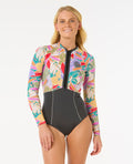 Mujer con traje de neopreno RIP CURL G-BOMB 1.5MM FZ SPRINGSUIT de manga larga y cremallera frontal, con estampado tropical y parte inferior negra, posando de frente.