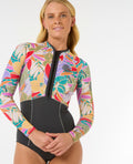 Mujer con traje de neopreno RIP CURL G-BOMB 1.5MM FZ SPRINGSUIT de manga larga, con diseño tropical y cremallera frontal, sonriendo y en pose relajada.