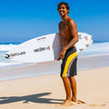 Bañador surf Rip Curl Mirage Archive amarillo y negro, foto lifestyle en playa con surfista y tabla Rip Curl, paneles cut-and-sew amarillo y piping gris.