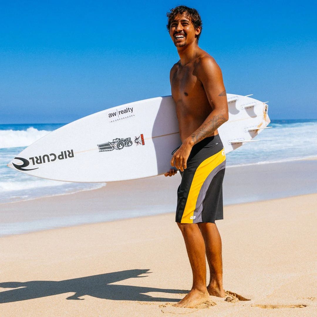 Bañador surf Rip Curl Mirage Archive amarillo y negro, foto lifestyle en playa con surfista y tabla Rip Curl, paneles cut-and-sew amarillo y piping gris.