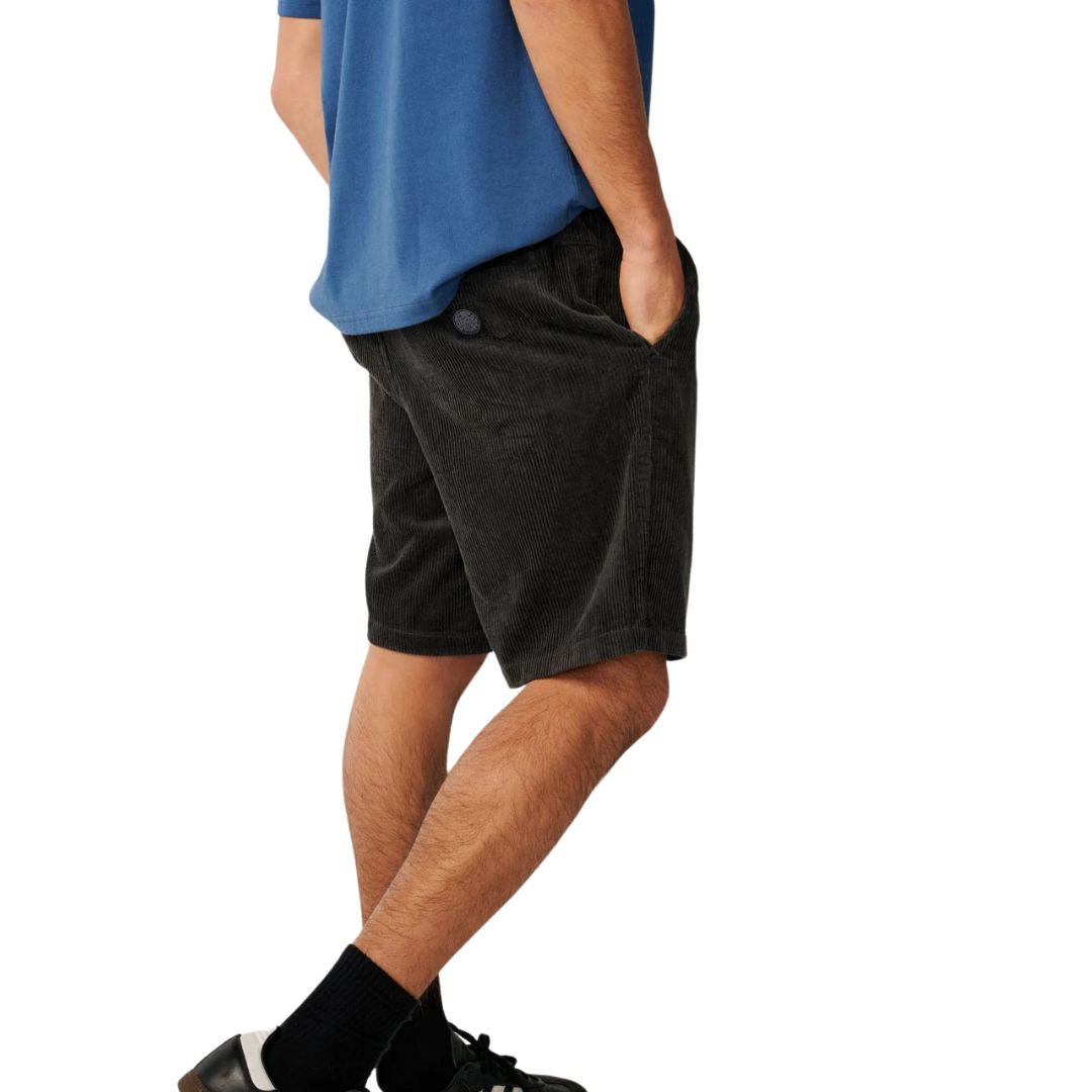 Pantalón corto pana Rip Curl Premium Surf Cord negro, vista lateral con camiseta azul, mano en bolsillo y logo bordado circular en pana acanalada acid wash.