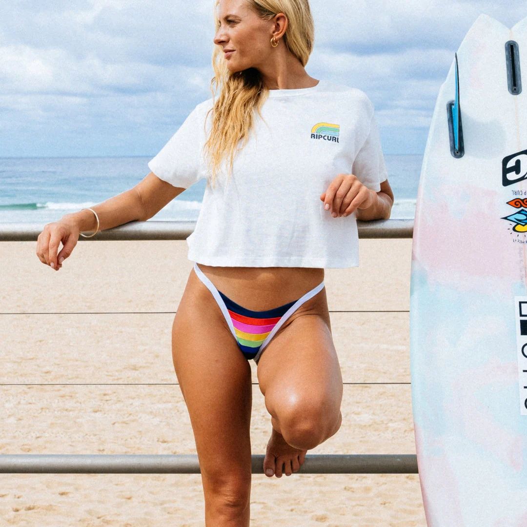 Surfista con braguita bikini Rip Curl Surf Stripe Cheeky dark navy rayas arcoíris y ribete blanco en paseo marítimo junto a tabla de surf.