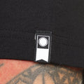 Detalle de la etiqueta en el brazo de la camiseta Salty Crew Kraken Monster en negro, con el logo circular de la marca cosido en el bajo, junto al dobladillo.