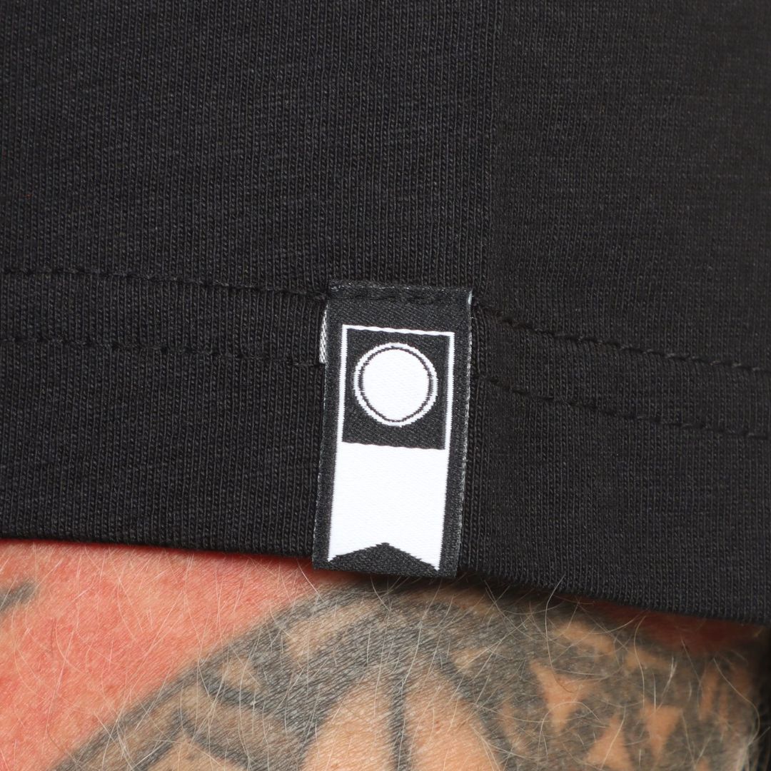 Detalle de la etiqueta en el brazo de la camiseta Salty Crew Kraken Monster en negro, con el logo circular de la marca cosido en el bajo, junto al dobladillo.