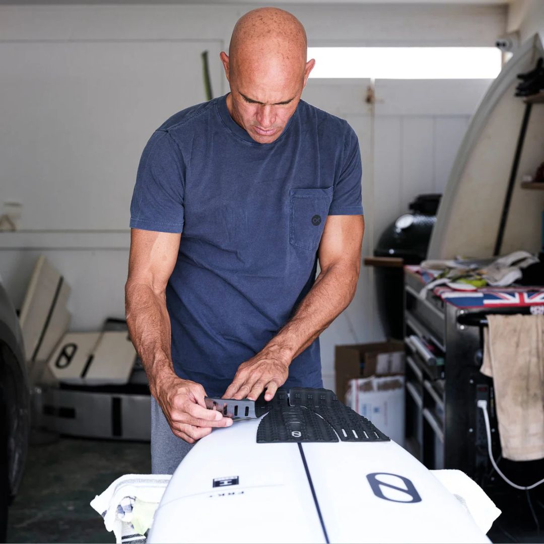 Kelly Slater colocando el grip trasero negro Slater Designs 3 piezas sobre una tabla de surf Firewire blanca en su taller, ajustando la pieza central del arco.