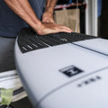 Detalle de manos presionando el grip de surf trasero Slater Designs negro sobre tabla Firewire gris, adhesivo 3M en proceso de fijación, textura cuadrada BLOOM visible de cerca.