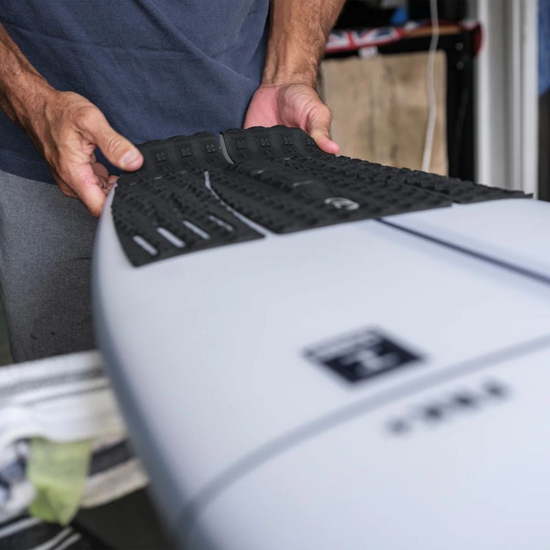 Kelly Slater posicionando las piezas del grip trasero negro Slater Designs sobre tabla Firewire gris, mostrando la instalación por secciones del pad de surf de 3 piezas con arco.