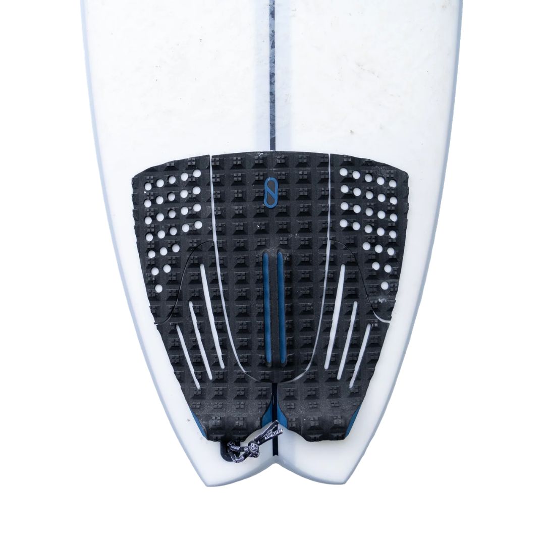 Grip Slater Designs 5 Piece Action Pad negro y azul instalado en tabla de surf Firewire blanca, vista cenital de la cola mostrando las 5 piezas, arco bajo azul, ranuras paralelas y recortes circulares.