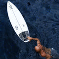Vista aérea de Kelly Slater en el agua junto a su tabla Slater Designs FRK+ blanca, con el grip trasero 5 Piece Action Pad negro instalado en la cola, remando en aguas azules profundas.
