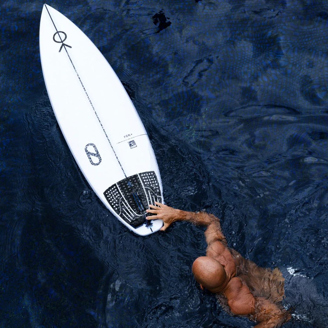 Vista aérea de Kelly Slater en el agua junto a su tabla Slater Designs FRK+ blanca, con el grip trasero 5 Piece Action Pad negro instalado en la cola, remando en aguas azules profundas.